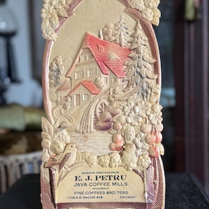 May include: An antique, beige-colored wall plaque with a relief carving of a house with a red roof, trees, and a watermill. The plaque has floral and bird details, and text that reads "E. J. Petru Java Coffee Mills, Fine Coffees and Teas, Chicago."
