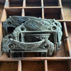 May include: A collection of dark gray metal drawer pulls featuring a curved, ornate design with two screw holes on each side for mounting. The hardware has a textured, weathered finish and is stacked together in a grid-like wooden compartment.