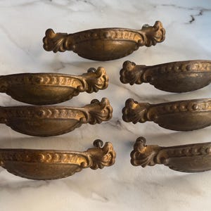 May include: Seven antique bronze drawer pulls with ornate, scroll-like detailing. The pulls have a curved, oval shape with a central, raised section and decorative accents. The hardware is a warm, aged bronze color, suggesting vintage charm.