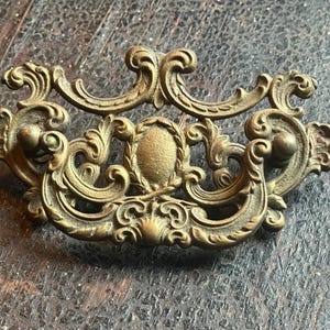 May include: Ornate brass drawer pull with a detailed, symmetrical design. The antique hardware features scrolling flourishes, a central oval, and a warm, aged patina. A decorative element for furniture.
