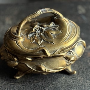 May include: A small, antique brass trinket box with a floral design on the lid and sides. The box has a handle and rests on three small feet. The brass has a warm, aged patina, giving it a vintage look.
