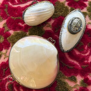 May include: Three iridescent shell-shaped purses with metal frames. The purses are oval and round, with a pearlescent sheen. They rest on a red and green floral patterned fabric.