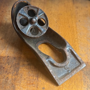 May include: A vintage, cast iron tool with a small wheel and a curved handle. The tool is a rusty brown color and appears to be an antique. The wheel has five circular openings. The handle has a curved opening and the letter "S".