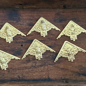 May include: Six gold-colored, triangular decorative corner pieces with a detailed face design. The pieces are arranged on a dark wooden surface, showcasing their intricate details and vintage aesthetic. Each corner piece features a central face with ornate headdress.