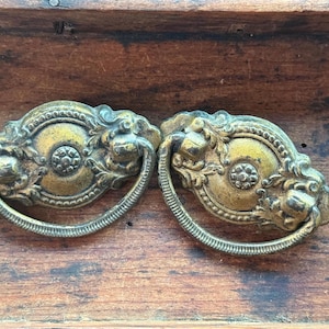 May include: Two oval-shaped brass drawer pulls featuring ornate, embossed floral and scrollwork detailing. Each piece includes a central circular medallion with a small flower motif and a swinging, ribbed semi-circular handle attached to the backplate.