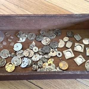 May include: A rectangular wooden box containing a collection of assorted vintage watch parts, including circular metal movements, metallic watch cases, small clear glass crystals, and various tiny mechanical components scattered across the base.