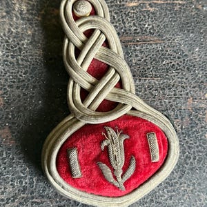 May include: Vintage military shoulder board with a red velvet base and intricate silver-colored woven detailing. The design features a floral emblem and two rectangular bars, with a small button at the top. The overall aesthetic is ornate and historical.