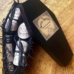 以下が含まれることがあります： 「Super-Natural Bathing Experience」と書かれたラベルが付いた黒い棺型の箱。箱が開いており、ボディオイル、石鹸、シャワージェルの入ったバス用品セットが入っています。商品は黒い薄紙に包まれ、黒いリボンで結ばれています。