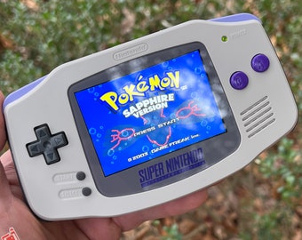 May include: A handheld Nintendo Game Boy Advance in a gray and purple color scheme. The screen displays the "Pokémon Sapphire Version" game. The device has a directional pad, start and select buttons, and purple A and B buttons.
