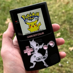 May include: A black Game Boy Color handheld gaming console with a pink and white custom design featuring a Pokemon character. The screen displays the Pokemon Yellow game with Pikachu on the screen. The text "GAME BOY COLOR" is printed on the front of the console.