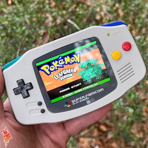 Puede incluir: Una consola de juegos portátil Game Boy Advance SP blanca y azul con una pantalla verde que muestra la pantalla de título de la versión Pokemon LeafGreen. La consola tiene un botón amarillo y rojo en el lado derecho y un botón de encendido en la parte superior.