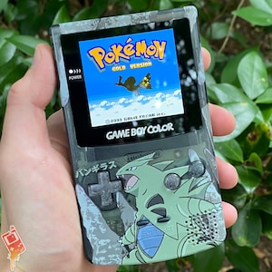Könnte beinhalten: Eine graue Game Boy Color Handheld-Spielkonsole mit Tarnmuster. Der Bildschirm zeigt das Pokemon Gold Version Spiel mit dem Text "Pokemon Gold Version" und "2000 GAME FREAK inc.". Die Konsole hat auch den Text "GAME BOY COLOR" und "バンギラス" aufgedruckt.