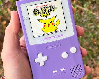 May include: A purple Game Boy Color handheld gaming console with a white screen displaying the Pokemon game. The game title is in Japanese and features a yellow Pikachu character. The console has white buttons and a speaker.