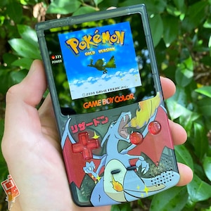 May include: A black Game Boy Color handheld gaming console with a red, white, and blue Pokemon Gold Version game cartridge inserted. The screen displays the Pokemon logo and the text "GOLD VERSION". The console has a red and black design with a graphic of a Charizard Pokemon.