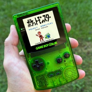 Puede incluir: Una consola de videojuegos portátil Game Boy Color verde con una carcasa verde translúcida. La pantalla muestra la pantalla de título del juego "Pocket Monsters" en japonés e inglés. La consola cuenta con un pad direccional negro, dos botones negros y un botón de encendido verde.