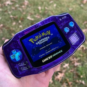 Puede incluir: Una consola de juegos portátil Game Boy Advance de color morado con una carcasa de plástico transparente. La pantalla muestra la pantalla de título del juego Pokemon Sapphire Version. El texto en la pantalla dice "Pokemon Sapphire Version" y "Presiona Start". El texto en la consola dice "Game Boy Advance".