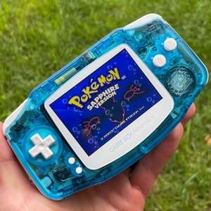May include: A blue and white Game Boy Advance SP handheld gaming console with a transparent blue shell. The screen displays the Pokemon Sapphire Version game.