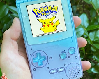 May include: A light blue Game Boy Color handheld gaming console with a translucent case. The screen displays the Pokemon Yellow game with Pikachu on the screen. The text on the screen reads "Pokemon Yellow Version" and "1995-1999 GAME FREAK Inc."