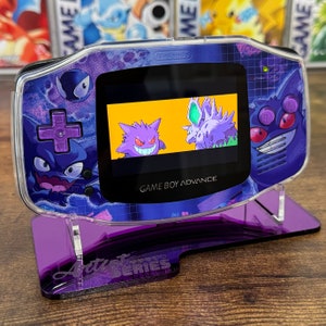 May include: A clear purple and blue Game Boy Advance with a purple and black stand. The screen shows a video game with three purple and black Pokemon characters. The stand has the text "Artist Series" on it.