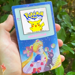 Puede incluir: Una consola de juegos portátil Game Boy Color azul con un diseño personalizado que presenta un personaje de Sailor Moon y un Pikachu de la serie de videojuegos Pokémon. La pantalla muestra el título del juego Pokémon Amarillo.
