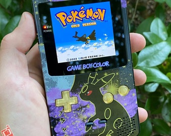 May include: A black Game Boy Color handheld gaming console with a custom design featuring a purple and black background with gold accents. The screen displays the Pokemon Gold game title and a black and white image of a Pokemon character. The text "Pokémon Gold Version" and "GAME BOY COLOR" are visible on the console.