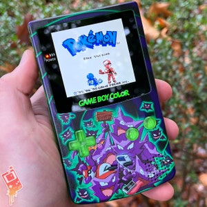 May include: A black and green Game Boy Color handheld gaming console with a custom design featuring a purple and green Gengar Pokemon character. The screen displays the Pokemon Blue Version title screen.