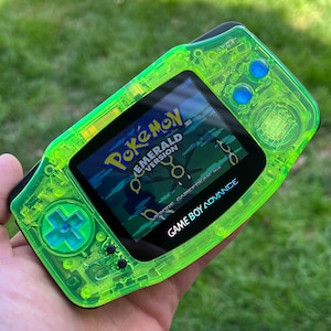 May include: A translucent green Game Boy Advance SP with a clear shell. The screen displays the Pokemon Emerald Version game title.