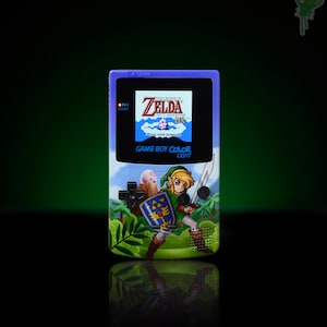 May include: A purple Game Boy Color Light handheld gaming console with a custom design featuring Link from The Legend of Zelda: Link's Awakening DX. The console has a green background with a forest scene and Link is holding a sword and shield.