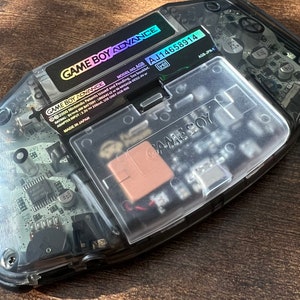 Könnte beinhalten: Eine transparente Handheld-Spielkonsole Game Boy Advance mit einem schwarzen und silbernen Etikett mit der Aufschrift "GAME BOY ADVANCE MODEL NO. AGB AJ14658914". Das Etikett enthält auch den Text "2000 Nintendo Patents issued and Pending, See booklet. RATING: V0.6W BATTERY: LRSIAAN-MAGB-00332.4V or ADAPTER INPUT: 3.3V 350mA. USE ONLY AGS-008 MADE IN JAPAN AGB-JPN-1".
