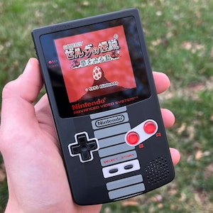 Puede incluir: Una consola de videojuegos portátil Game Boy Color negra con una disposición de botones rojos y blancos. La pantalla muestra el juego "The Legend of Zelda: Link's Awakening" en japonés. El texto "Nintendo" y "Advanced Video System" está impreso en la consola.