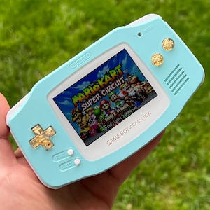 May include: A light blue Game Boy Advance handheld gaming console with a white faceplate. The console is displaying the game Mario Kart: Super Circuit. The console has gold accents on the buttons and the power button.