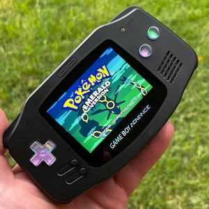 May include: A black Game Boy Advance handheld console displaying the "Pokémon Emerald Version" game. The screen shows a green and blue landscape with a Pokémon character. The console has a directional pad, start and select buttons, and power button.
