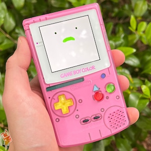 Puede incluir: Una consola de videojuegos portátil Game Boy Color rosa con una pantalla blanca que muestra una línea verde. La consola tiene un pad direccional amarillo, botones rojo y verde y un altavoz.