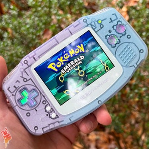 Könnte beinhalten: Ein Game Boy Advance SP in Lavendel und Blau mit glitzernder Oberfläche. Der Bildschirm zeigt das Spiel "Pokémon Smaragd Edition" mit dem Text "Game Freak Inc." und "Game Boy Advance".