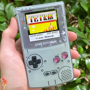 Puede incluir: Una consola de juegos portátil Game Boy Color gris con una carcasa de plástico transparente. La pantalla muestra el juego Tetris con el texto "DOT MATRIX WITH STEREO SOUND" y "TETRIS TM" en la parte superior. La pantalla del juego muestra el juego en progreso con el texto "1 PLAYER", "2 PLAYER", "©1989 Nintendo", y "GAME BOY COLOR".