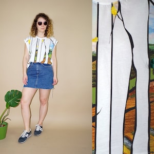 以下が含まれることがあります： カラフルな風景デザインの白いキャップスリーブTシャツとデニムミニスカート、スニーカーの組み合わせ。Tシャツには垂直の黒い線と、緑、黄色、茶色の色合いの風景イラストが描かれています。