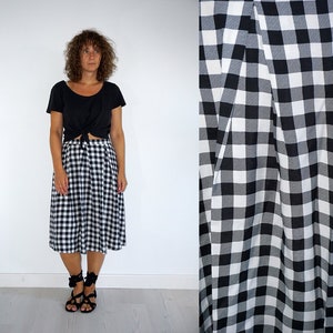 Pode incluir: Uma saia midi de xadrez vichy em preto e branco com cintura alta e design plissado. A saia é feita de um tecido leve e tem uma silhueta fluida.