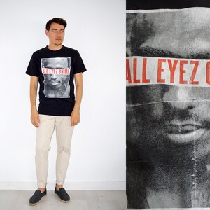 May include: Black t-shirt with a graphic of a man's face and the text "ALL EYEZ ON ME" in red. The shirt is paired with beige pants and dark shoes. The image is taken in front of a white background.