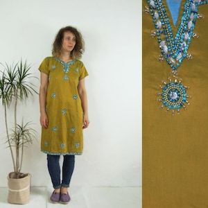 Puede incluir: Vestido túnica color mostaza con mangas cortas y un intrincado trabajo de cuentas turquesas y plateadas alrededor del escote y dispersas en la parte delantera. El vestido llega a la rodilla y se usa con jeans azules y zapatos morados. Una planta en maceta de yute a la izquierda.