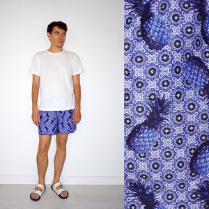 May include: A person wearing a white t-shirt, blue and white patterned shorts, and white sandals. The shorts feature a repeating pattern of blue pineapples on a blue and white geometric background. The image is set against a white backdrop.