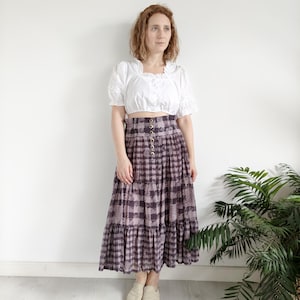 May include: A woman wearing a white, ruffled crop top and a long, tiered skirt with a black and white checked pattern. The skirt has a high waist and a button closure.
