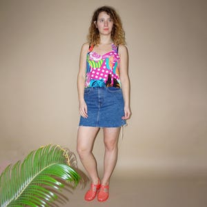 May include: A sleeveless top featuring a vibrant, multi-colored pattern of pink roses, white polka dots, and abstract shapes, paired with a medium-wash blue denim mini skirt with a raw hem and bright coral-colored jelly sandals.