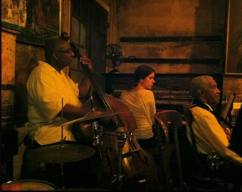 May include: A dimly lit stage with three musicians performing. A man in a yellow shirt plays a double bass, a woman in a white shirt stands to the right, and a man in a white shirt sits to the right of the stage. The stage is lit with warm, yellow light.