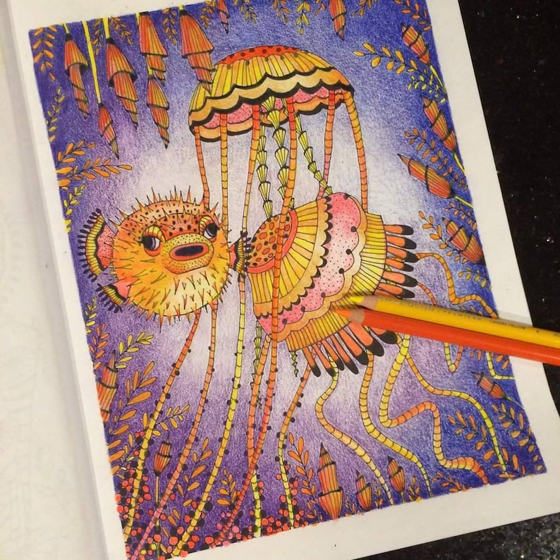 A completed coloring book page featuring a puffer fish and two jelly fish. Colored pencils used are sitting on top of the page. The picture is colored in oranges pinks and yellows on a purple background.