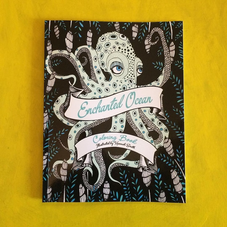The front cover of the Enchanted Ocean Coloring Book. An octopus illustration partially is colored in pale greens on a black background. The book is sitting on a yellow surface.