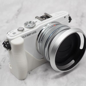 Puede incluir: Una cámara Olympus blanca con una lente plateada y una capucha de lente negra. La cámara tiene una correa negra.