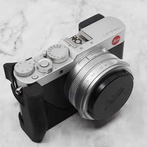 May include: A silver Leica camera with a black lens and a black leather grip. The camera has a black lens cap with the Leica logo.