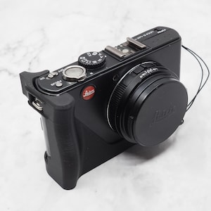Può includere: Una fotocamera digitale Leica nera con un copriobiettivo e una custodia protettiva nera. La fotocamera ha un logo Leica rosso e un piccolo flash in cima.
