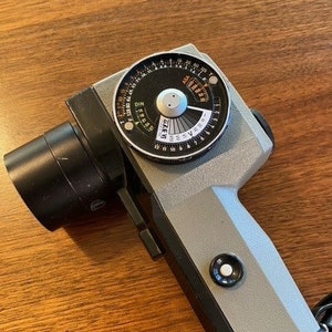 May include: A gray and black handheld light meter with a circular dial that reads "DIN" and "EV" and has numbers from 1 to 18. The dial is labeled with "ASA" and "25-50-100-200-400-800-1600".