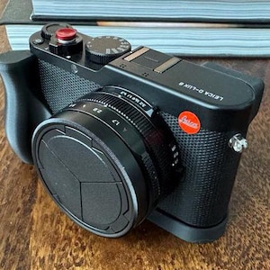 May include: A black Leica D-Lux 9 camera with a lens cap and a red button on the top. The camera has a textured black finish and a silver accent on the side.